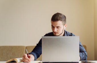 The Ultimate Guide to Finding a Reliable Thesis Writing Service Online A man ata table using a laptop with a pen beside him engaged in work or writing thesis.