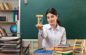 How to Manage Your Time During GCSE English Exams in Greenford? Female teacher in a classroom holding an hourglass, symbolizing time management, with books and study tools on the desk—ideal for conveying GCSE English exams time management tips for Greenford pupils.