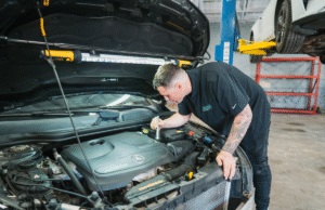 Top 5 Challenges for Independent Repair Shops in 2024 A mechanic inspects the engine of a Mercedes-Benz vehicle using a flashlight in an auto repair shop. The car’s hood is open with a work light attached, and another vehicle is visible on a hydraulic lift in the background.