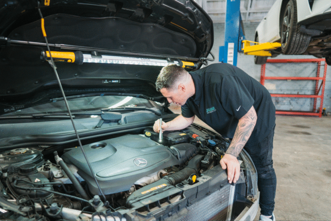 A mechanic inspects the engine of a Mercedes-Benz vehicle using a flashlight in an auto repair shop. The car’s hood is open with a work light attached, and another vehicle is visible on a hydraulic lift in the background.
