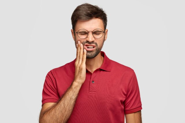 Photo of Caucasian man suffers painful toothache, has rotten tooth, needs to visit dentist, keeps eyes closed from pain, dressed in casual red t shirt, isolated over white background, touches cheek Weird tooth symptoms like numbness, bad taste, or painless tooth loss? Discover 8 strange dental issues you should never ignore—your health may depend on it.